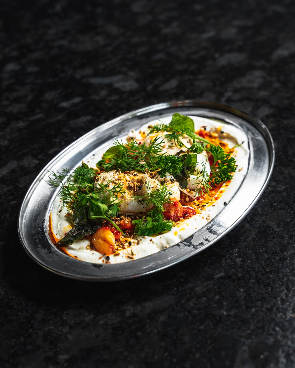 Eggs under fresh herbs on a creamy base with red chilli oil, on a steel oval plate