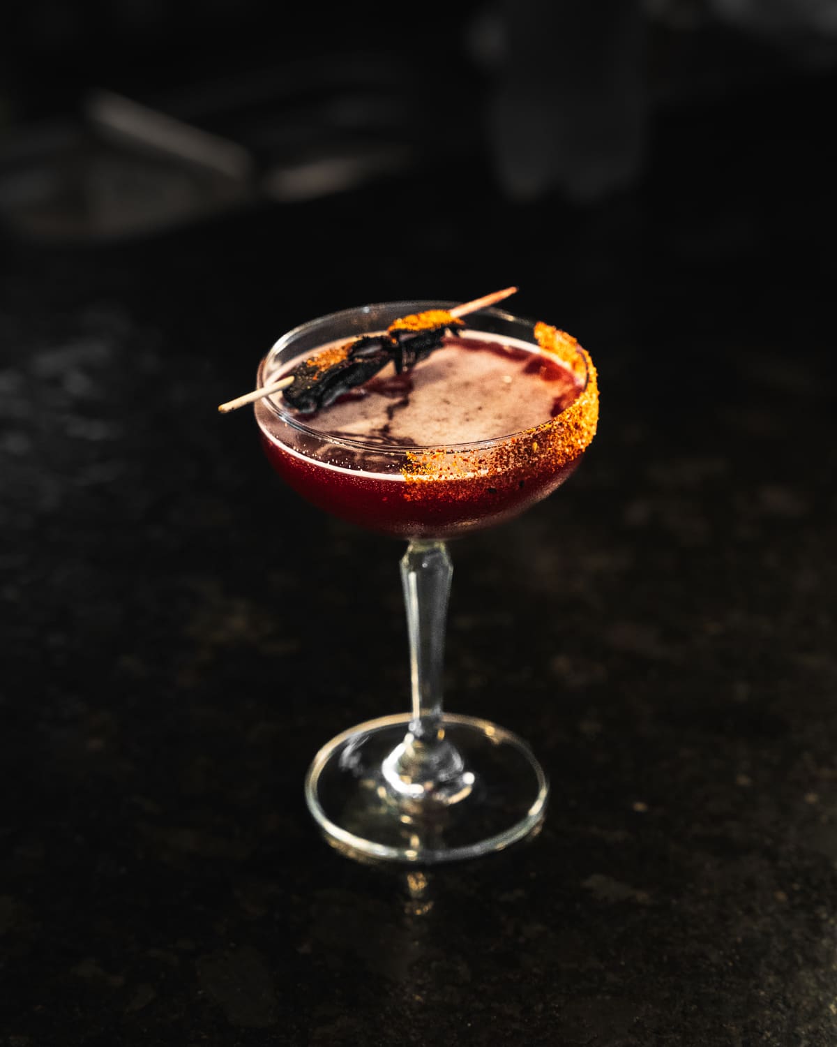 A red cocktail in a coupe with a spiced rim and a skewer garnish